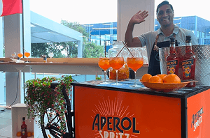A professional bartender waving from behind a fully customisable drinks tricycle featuring Aperol Spritz branding for a corporate summer party
