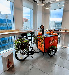 A professional bartender smiling and waving behind a branded drinks tricycle serving premium cocktails for a corporate summer event in a modern office space