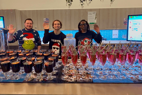 Three professional bartenders in festive jumpers smiling behind a mobile bar serving mulled wine and winter cocktails for an office staff party.